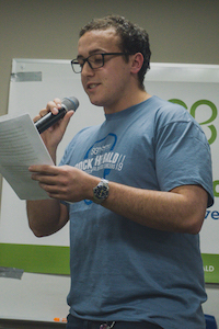 man in a blue shirt speaking into a microphone, holding a sheet of paper