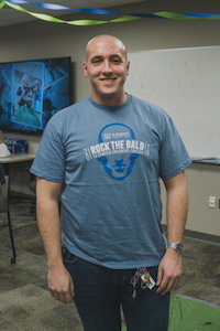 man with a shaved head in a blue t-shirt that says Rock the Bald, smiling at the camera