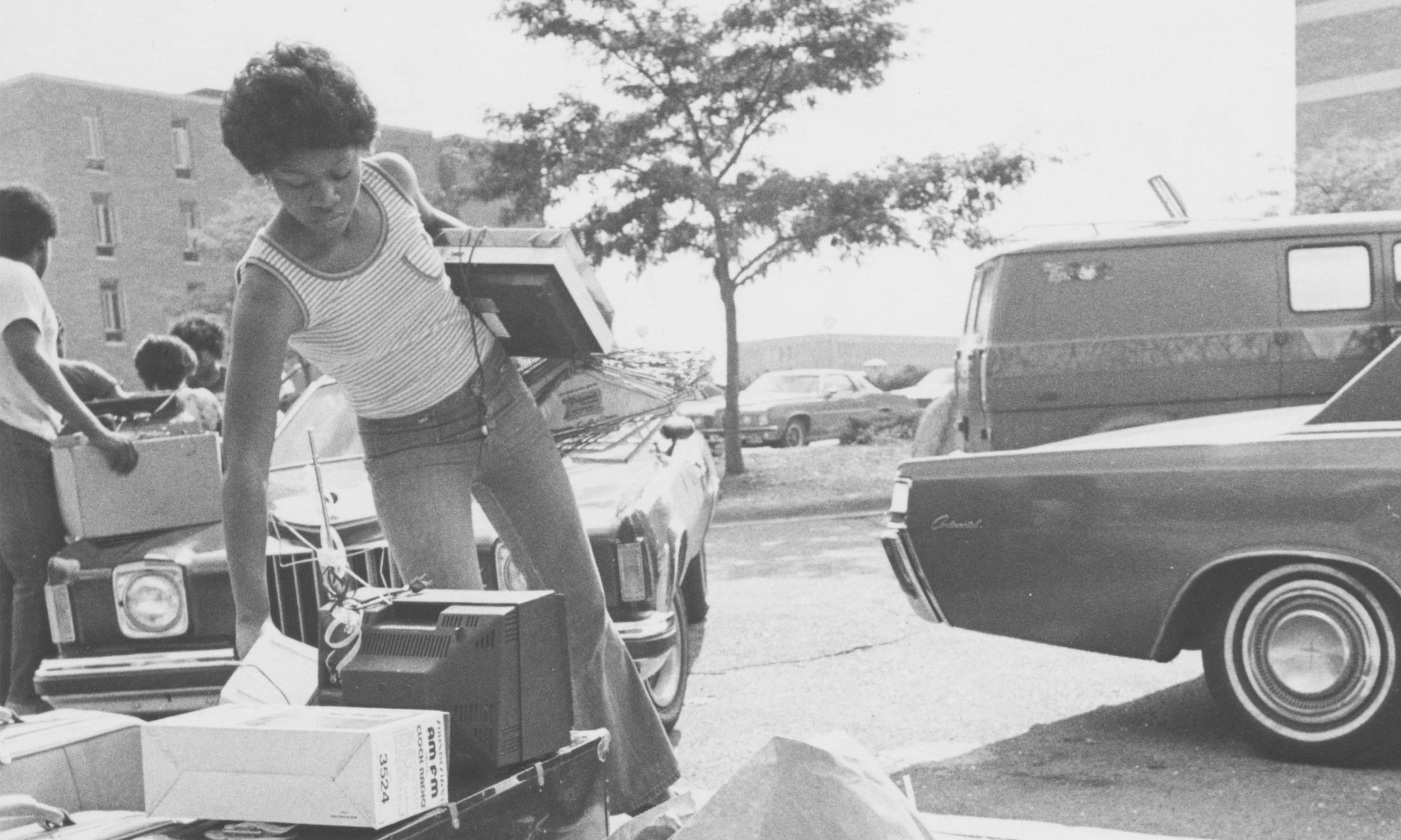 A black and white photo of an African-American Oakland University student picking up electronics from the curbside in front of an old styled car