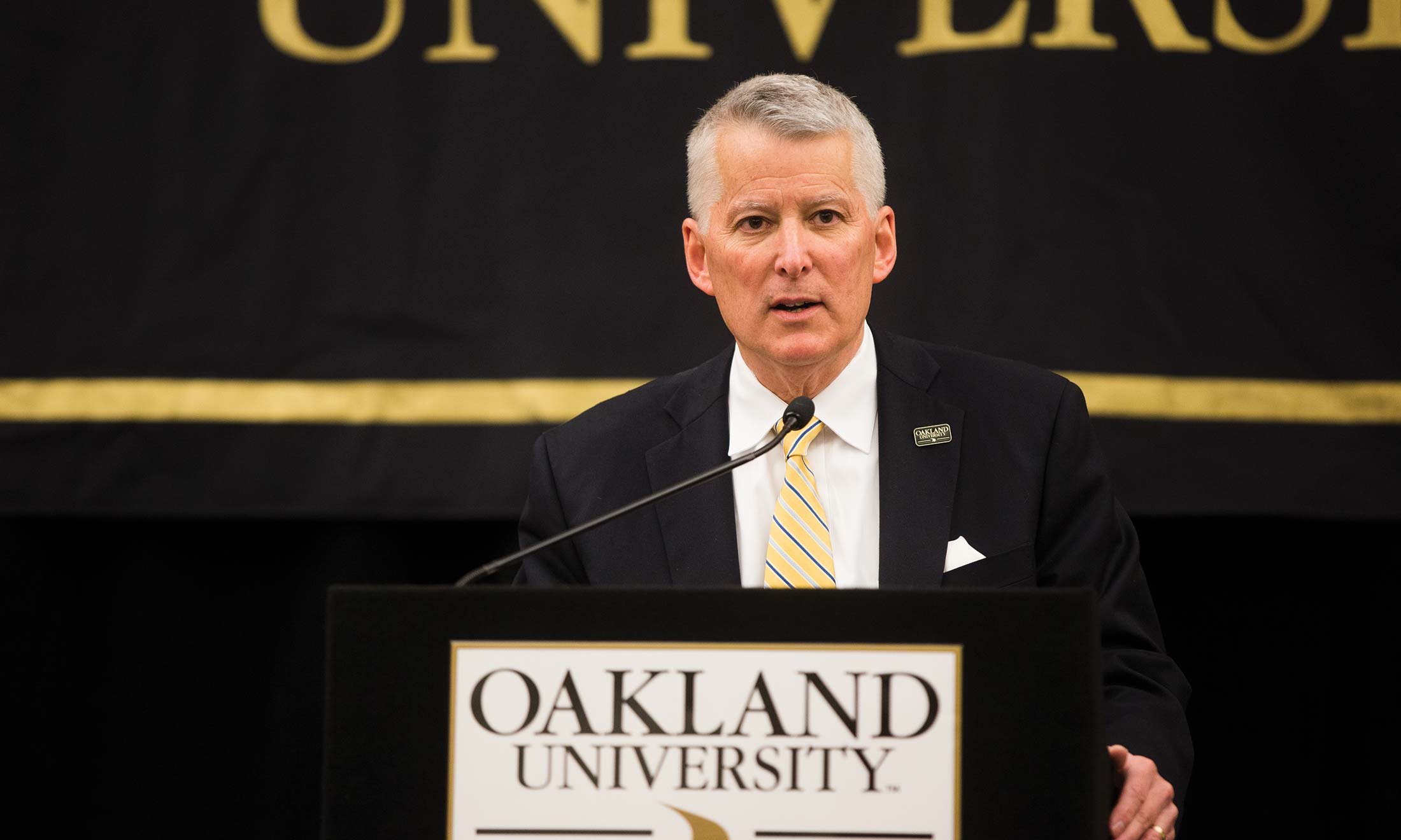 O U Board Chair Richard L. DeVore standing at podium announcing Dr. Ora Hirsch Pescovitz as the seventh president of Oakland University