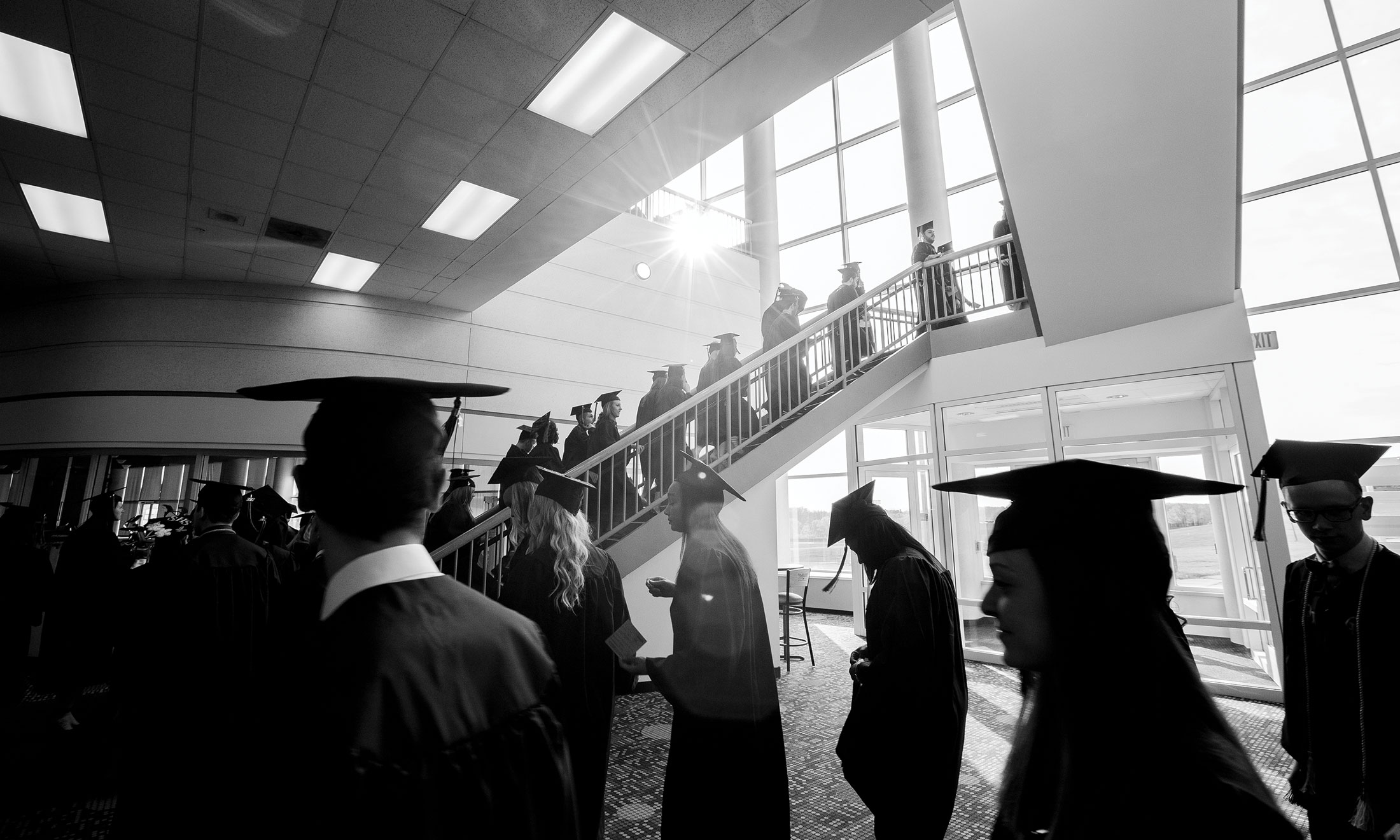 Black and white photo of students at their graduation, walking in a line up a staircase, wearing their caps and gowns