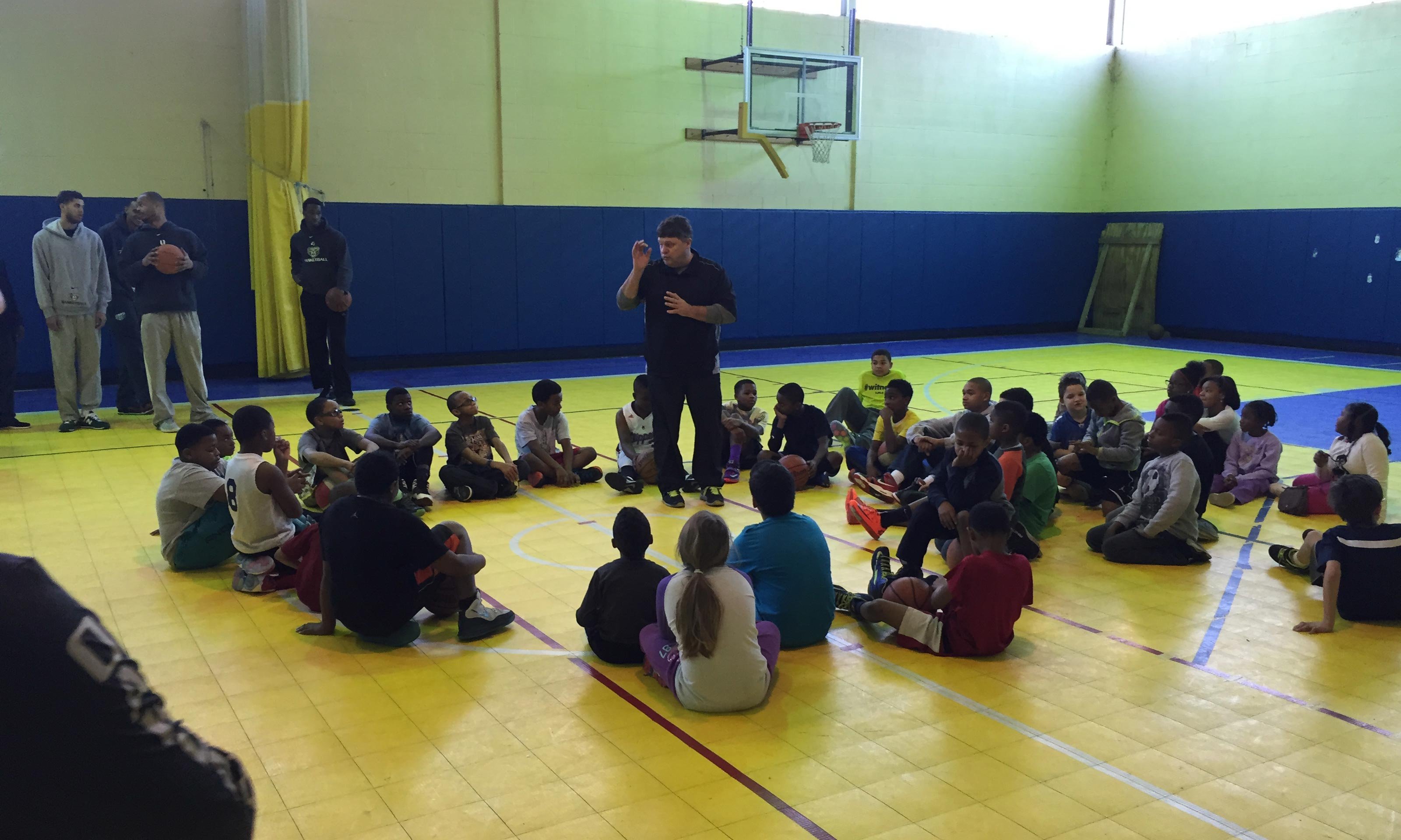 Coach Greg Kampe speaks to elementary kids seated circled around him in a gym.