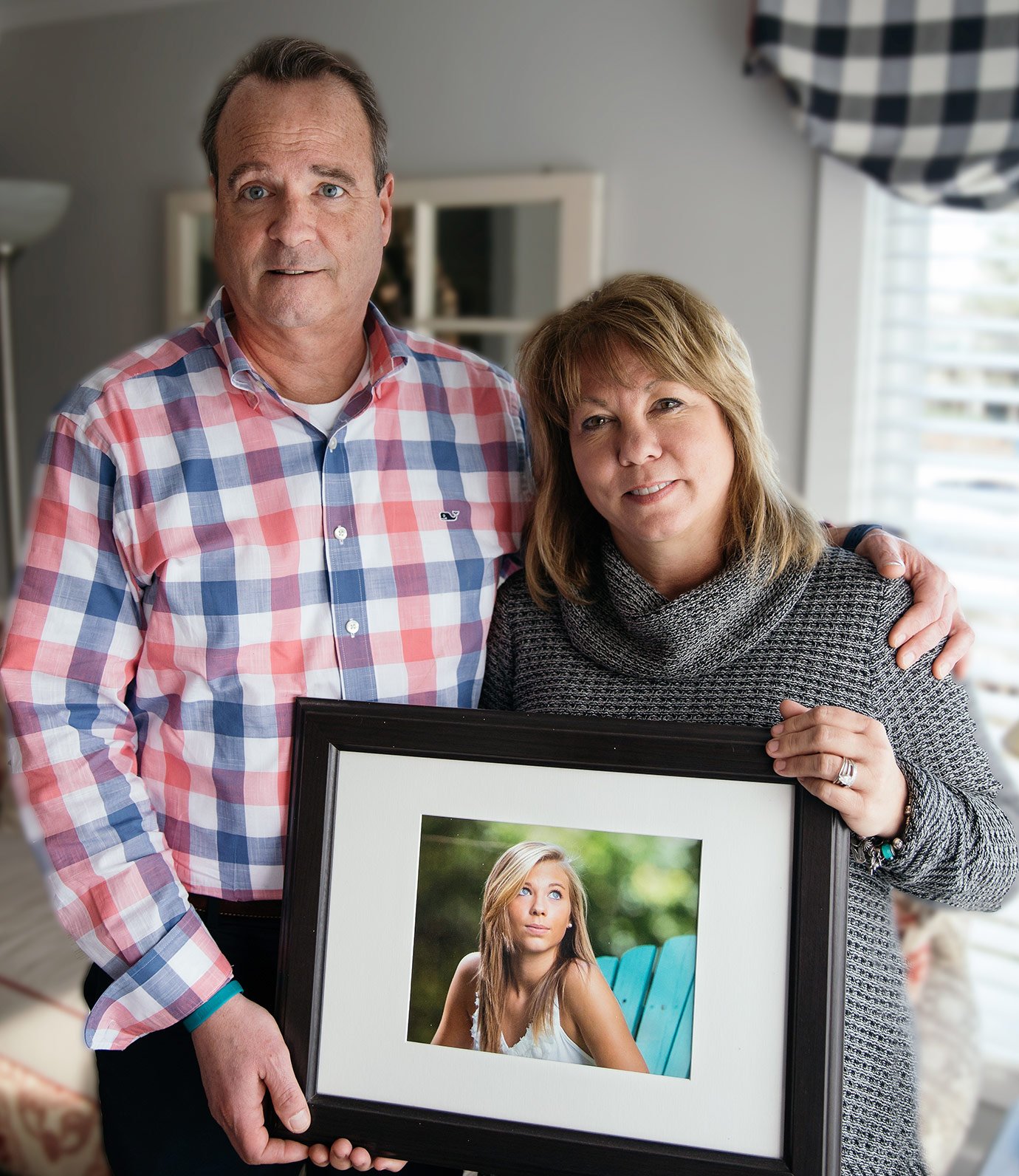 Wayne and Lori Brouilllet standing next to each, both holding onto a picture frame with a photo of their daughter in it. They're inside of a house standing in front of a window. 