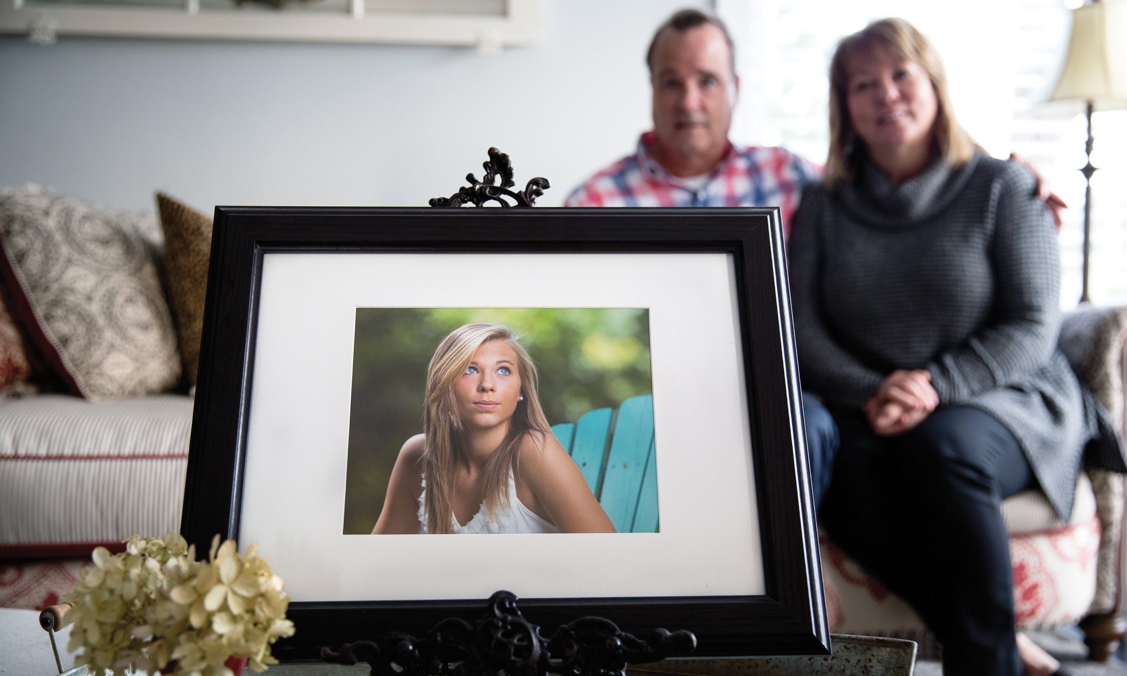 A picture in a frame of Hailey Brouillet in a teal wooden chair outdoors looking up at the sky. In the blurry background behind the picture, sits Wayne and Lori Brouilllet in their living room.