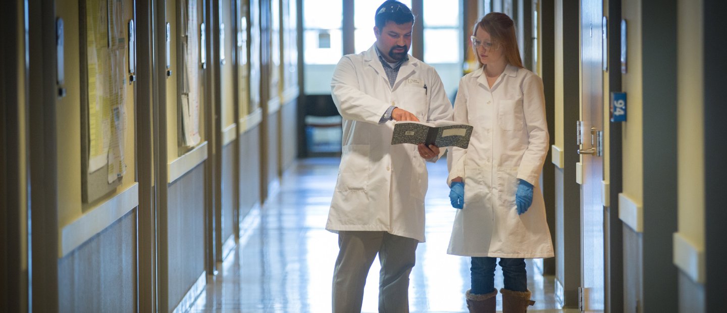 A man and a woman, wearing lab coats, standing in a hallway reading from a notebook.