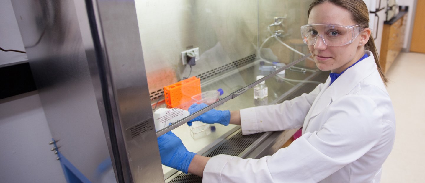 A woman in a lab coat, working with chemicals behind a glass shield.