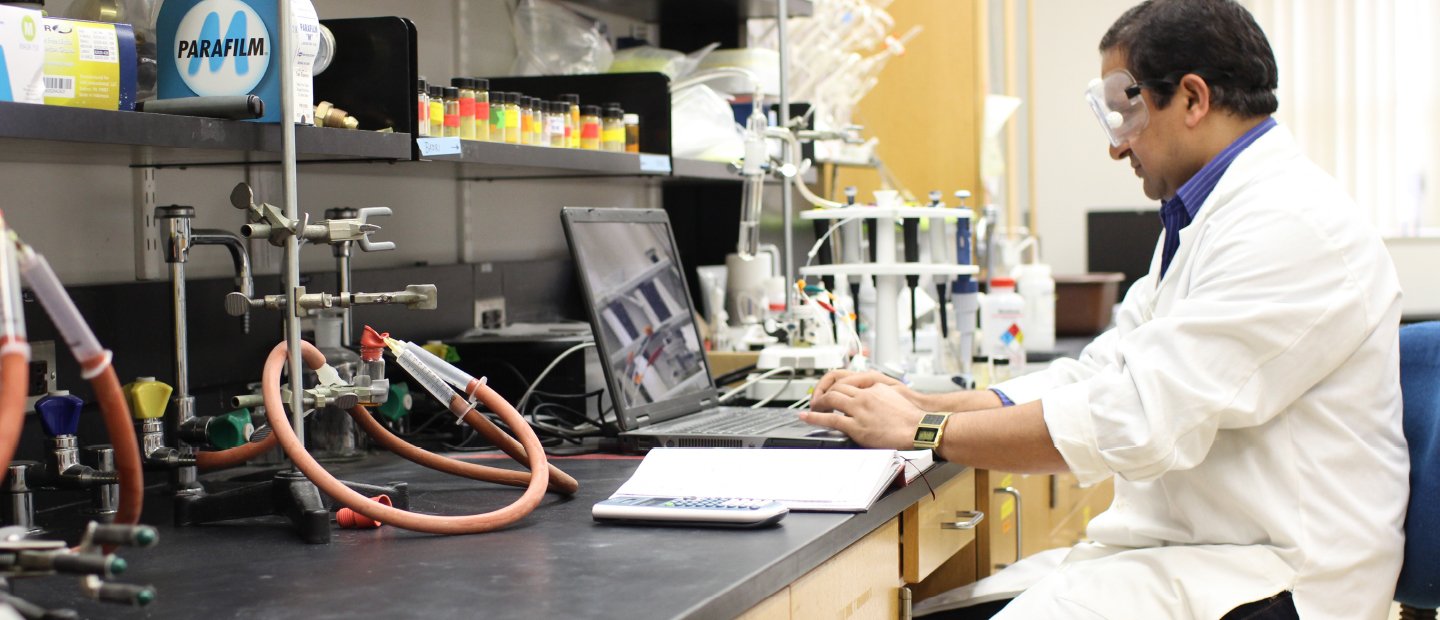 A man in a lab coat working on a laptop in a chemistry lab.