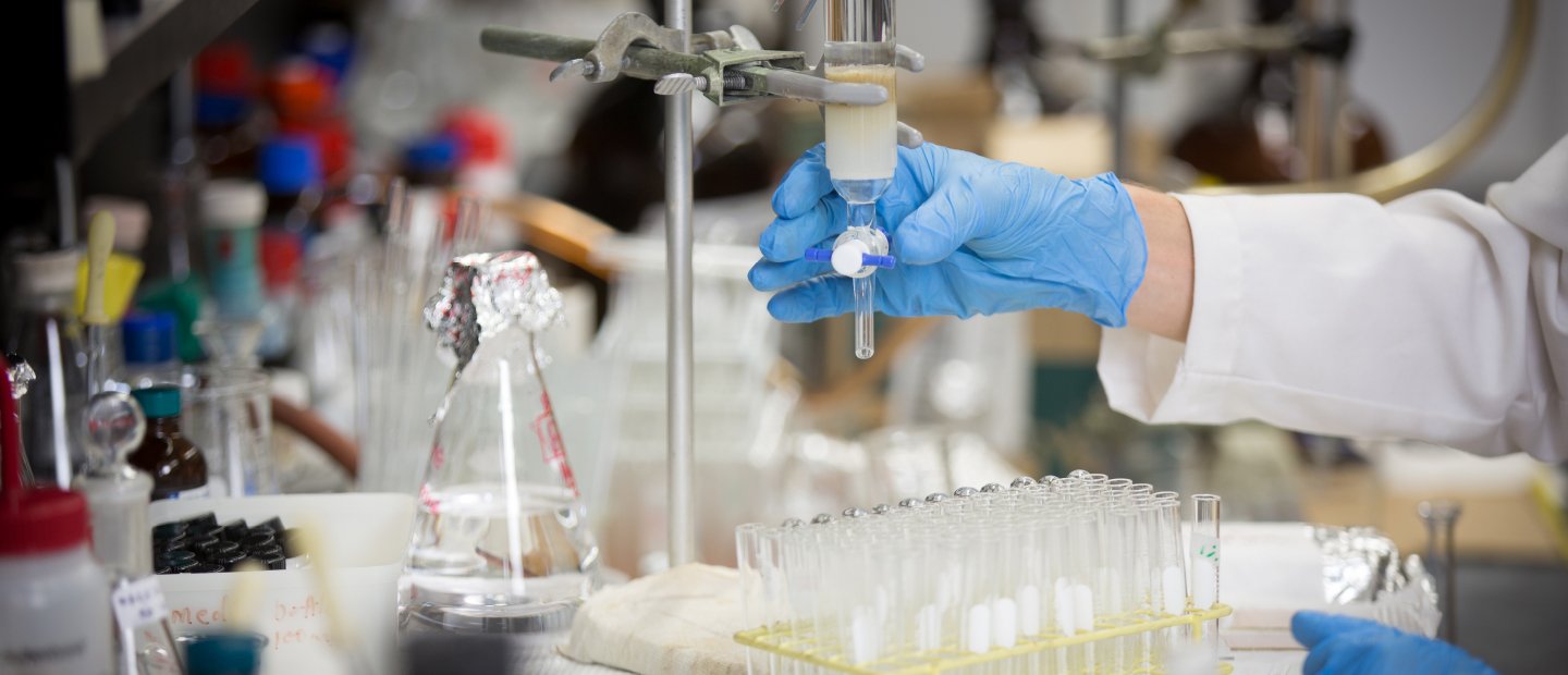 Research A gloved hand adjusting a valve on a glass vial, above a table with test tubes in a tray.