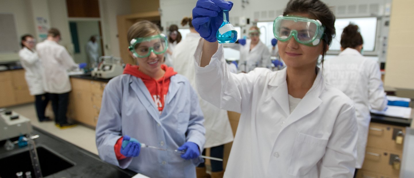Students wearing lab coats, holding glass beakers of chemicals, in a lab.