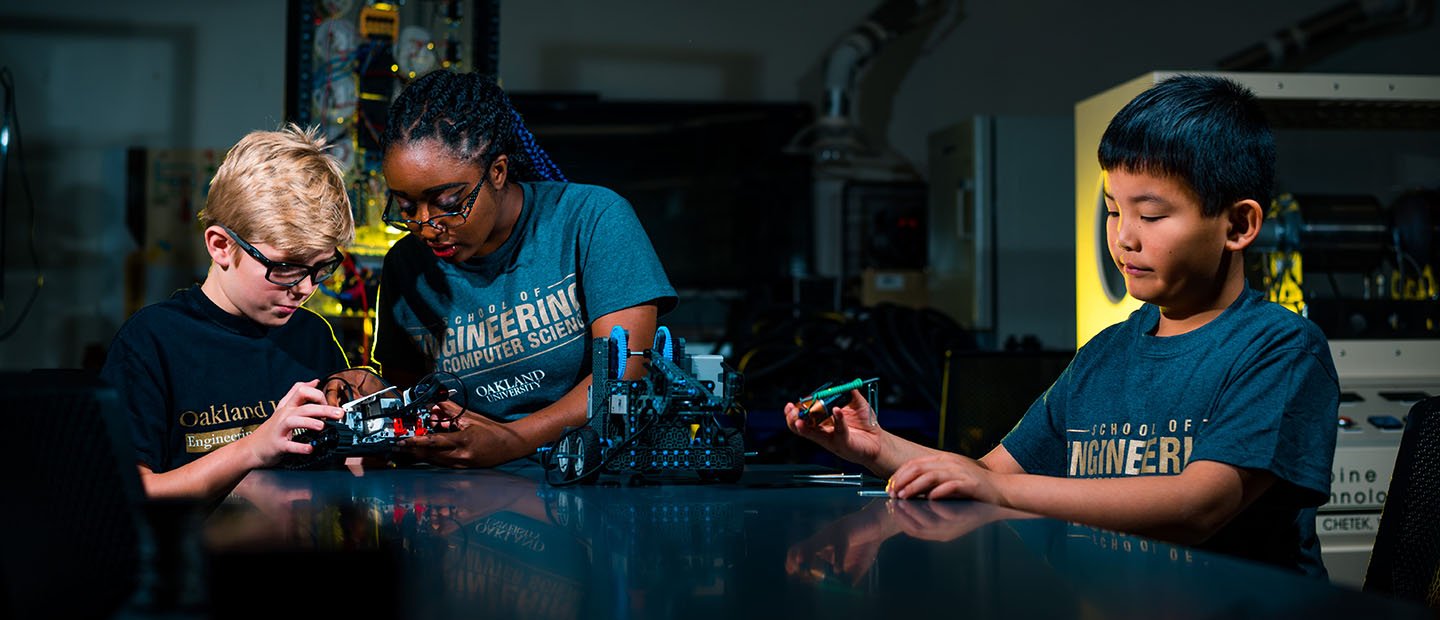 three kids standing around a table in a lab, working with mechanical equipment