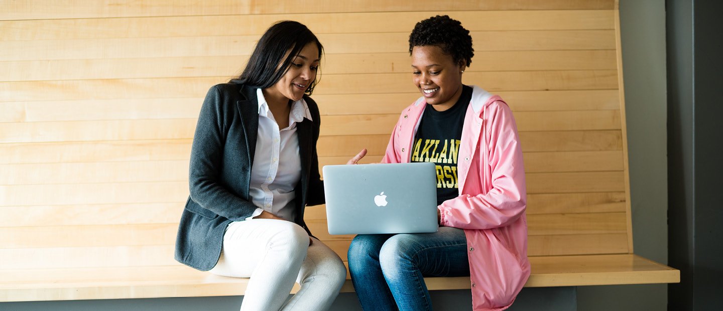 two young women seated on a wooden bench, looking at a laptop screen