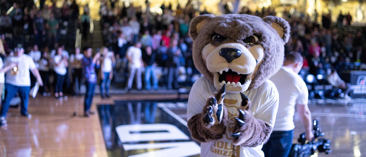 Homecoming Grizz bear mascot standing on an athletic court with people seated in the stands