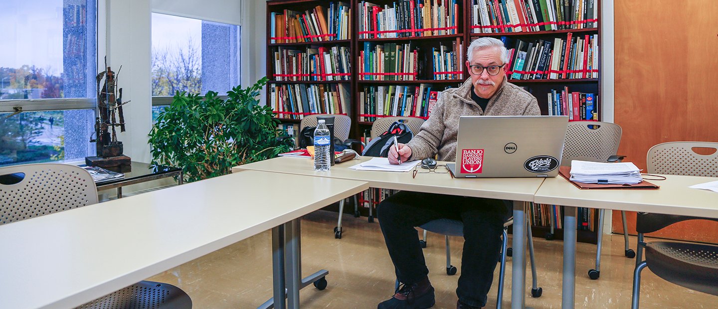 man seated at a table with paper and pen and a laptop, in front of a wall of book shelves