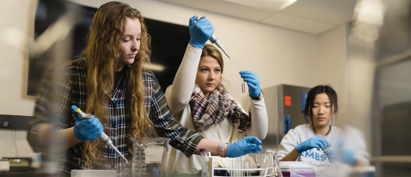 Three young women wearing blue gloves, working with test tubes and beakers in a lab.