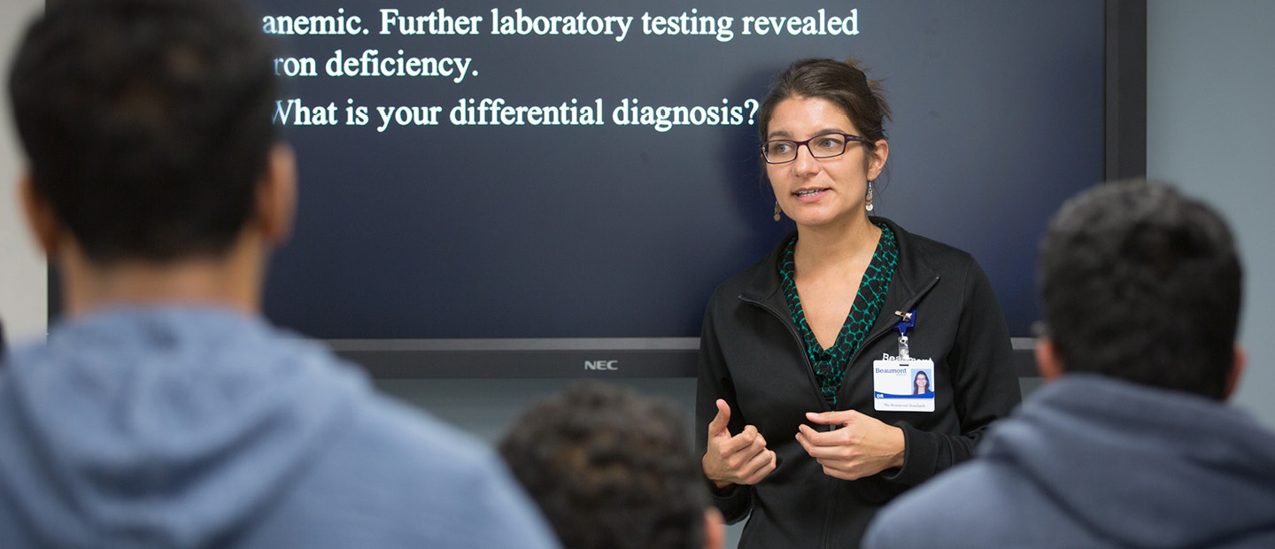 A woman wearing an ID badge, standing in front of a screen, presenting to a group.