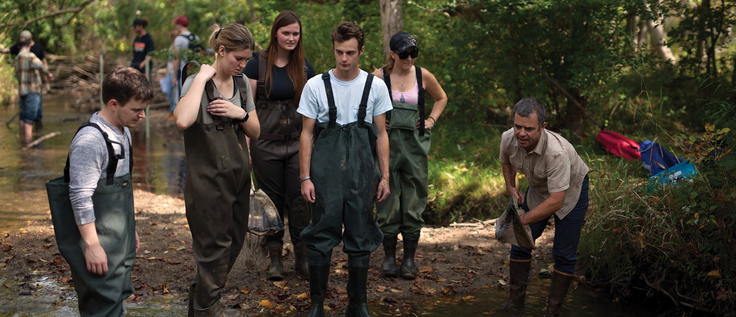 A group of students wearing overalls and boots, observing an instructor at the Biological Preserve.
