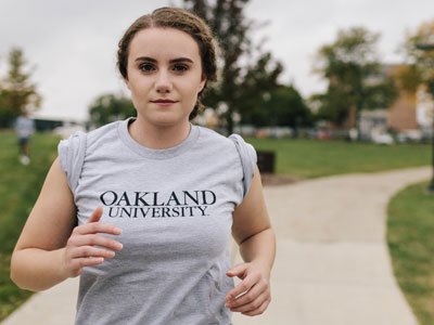 Woman wearing Oakland University tshirt, jogging on a sidewalk on campus