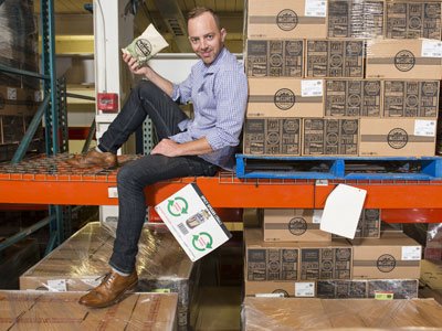 Man eating a bag of chips while sitting on a warehouse rack, leaning up against a pallet full of boxes.