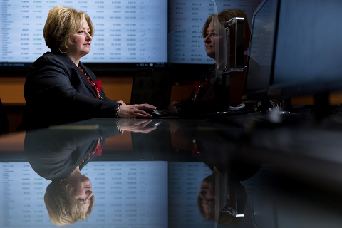Woman in a business suit sitting in a dark room with a screen of data behind her.