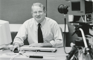 John Tower sits at a desk and is filmed with a video camera.