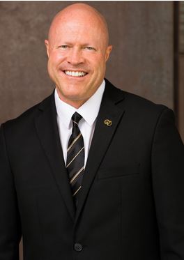 Male smiling for camera, dark jacket, tie, white shirt, OU pin on lapel.