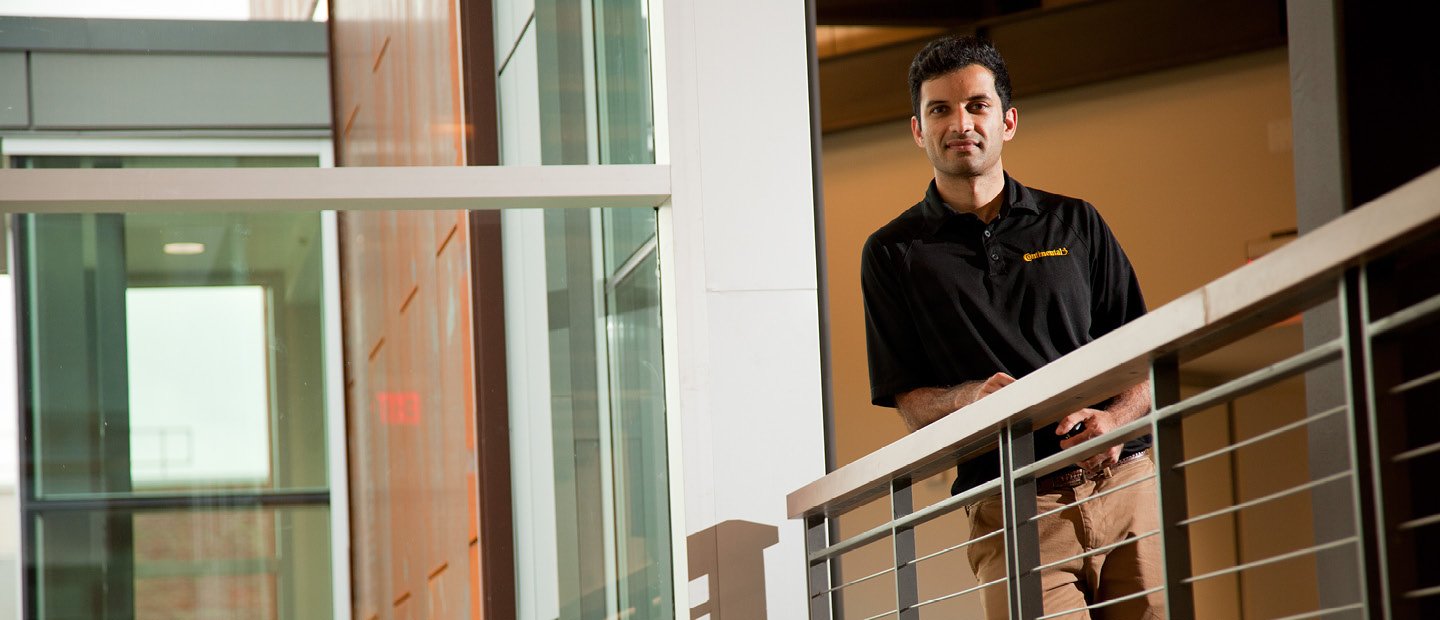 Business Essentials A man leaning on a railing in a hallway, looking down at the camera.