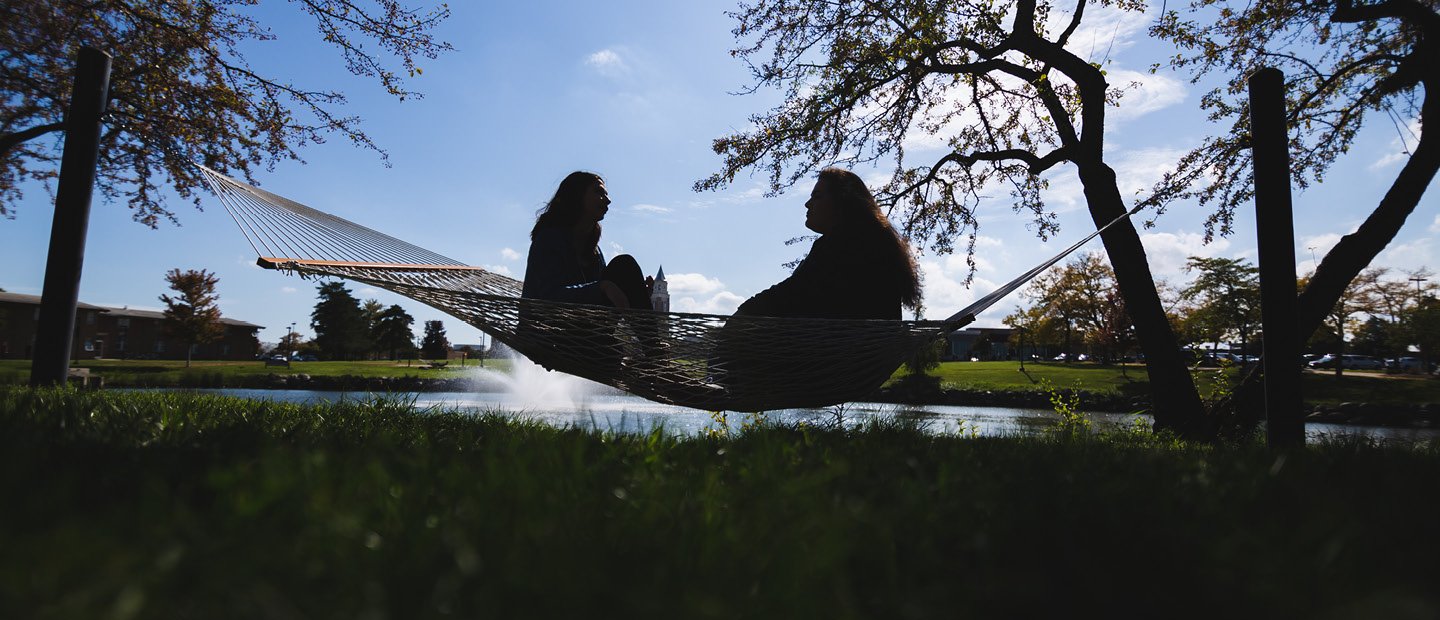 Two people seated, facing each other, in a hammock with a lake and fountain behind them.