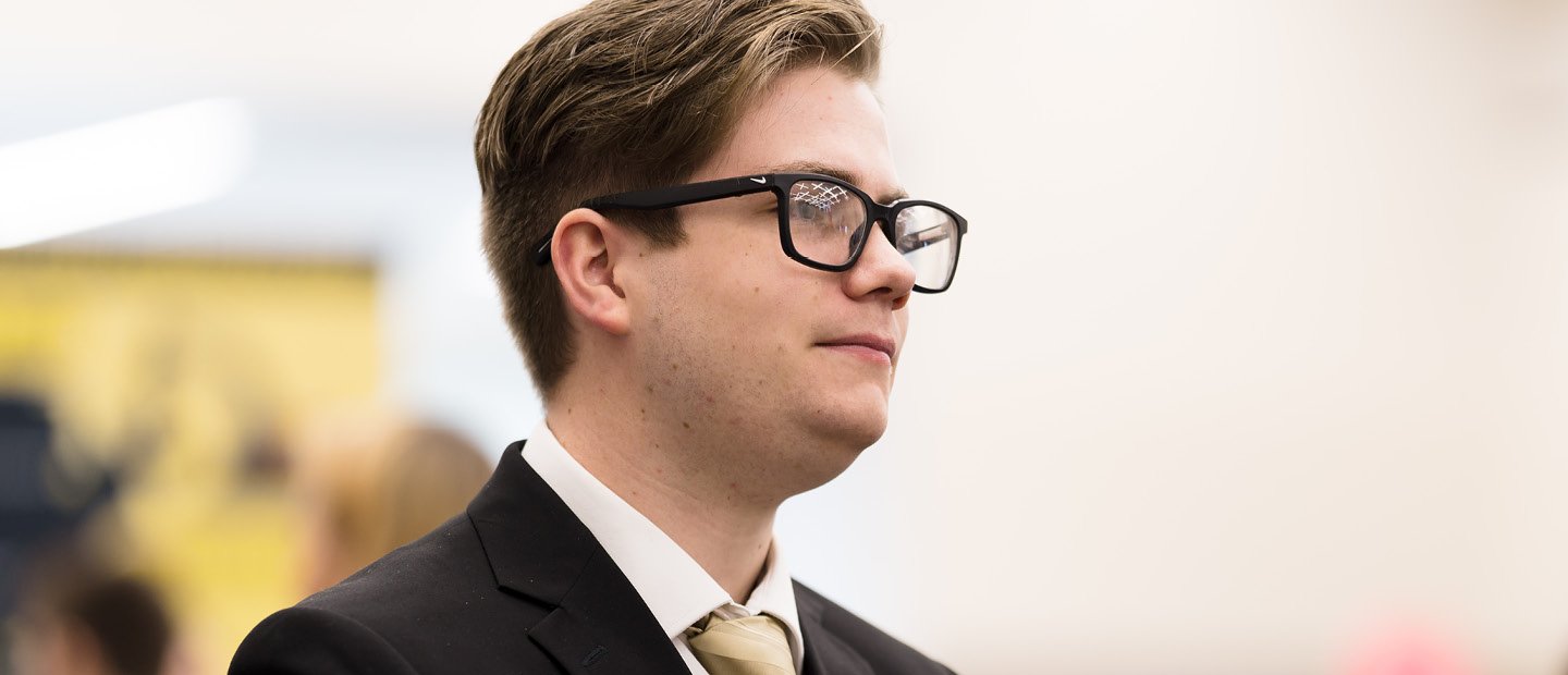 Economics A profile photo of a young man in glasses and a black suit jacket with a gold tie.