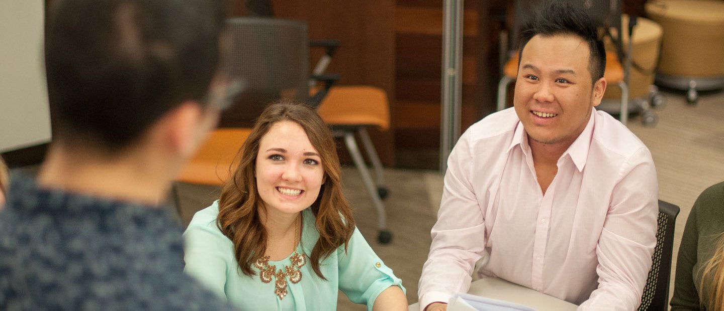 International Management Minor A man and woman seated at a desk, looking up and smiling at a man standing in front of them.