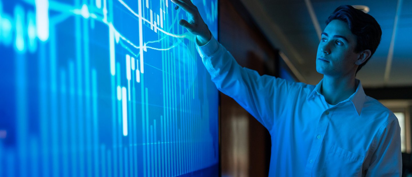 Undergraduate Students A young man pointing at a blue screen with a bar graph on it.
