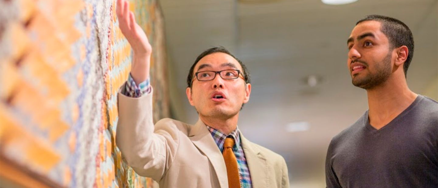 A professor gesturing to a colorful wall as a young man looks at it.