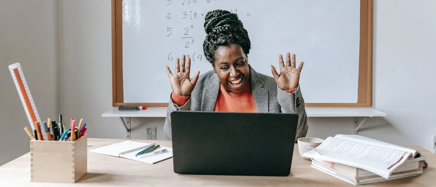 female teacher sitting at a desk in front of a laptop with her hands in the air