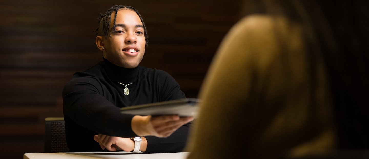 For Alumni A young man handing a portfolio to someone seated at a desk.