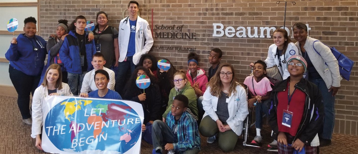 Group of adults and kids holding a sign with a picture of a world map that says "Let the Adventure Begin" 