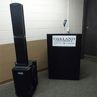 A black carpeted podium with Oakland University on the front, a wireless microphone, an Anchor Super PA speaker.