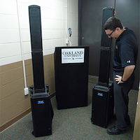 A black carpeted podium with Oakland University on the front, two wireless microphones, two Anchor Super PA speakers.