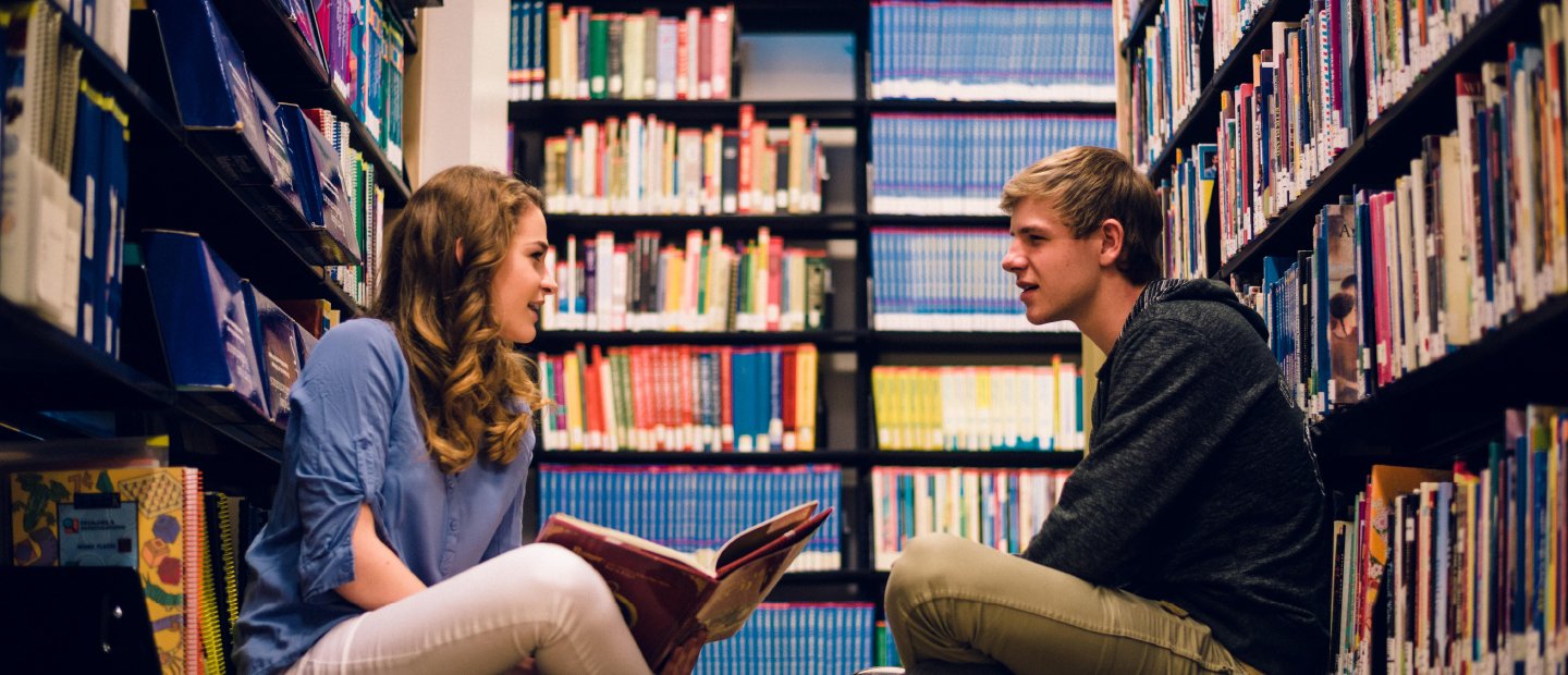 male and female student seated on the floor, reading, in a library