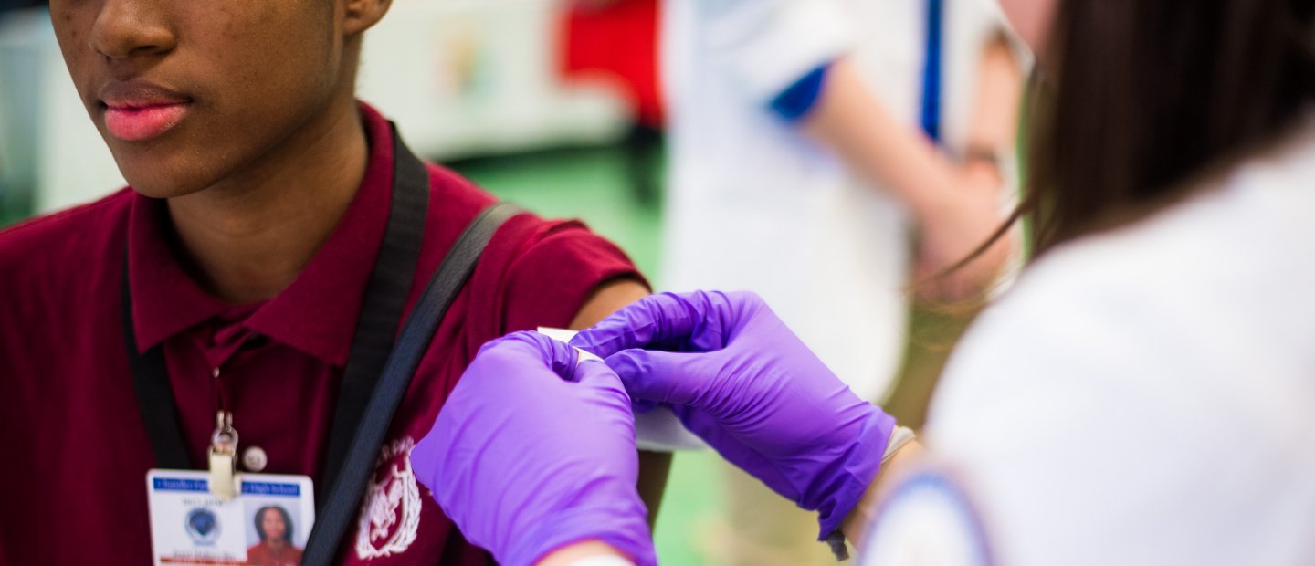 person in a white lab coat and purple gloves putting a band-aid on a student's arm