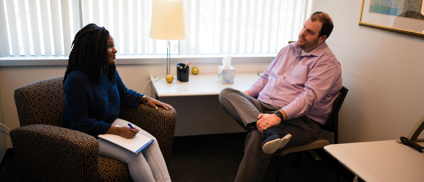Policies woman seated in an office, taking notes while talking to a man