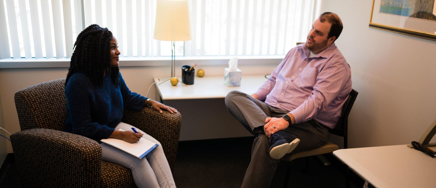 woman seated in an office, taking notes while talking to a man