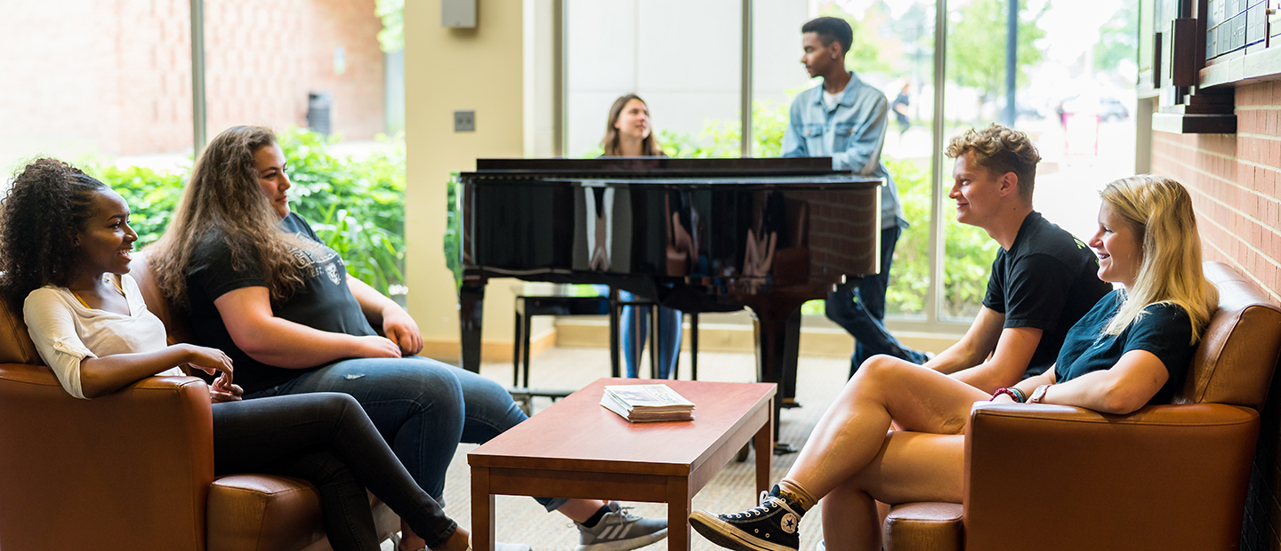 Two students at a piano while four students gather in a group in front of them.