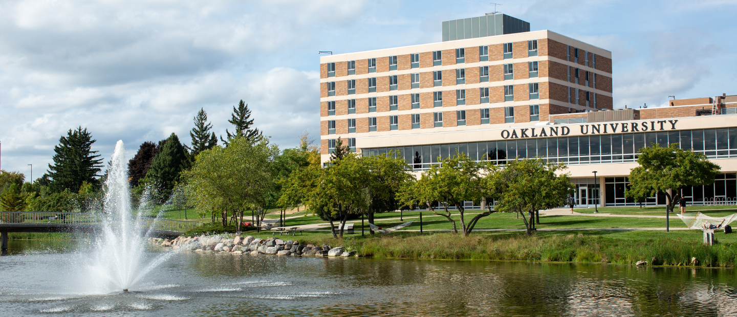 lake with a fountain, beige and brown building with Oakland University on it in the background