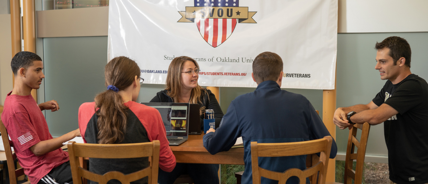 five people seated around a table with a red, white and blue banner behind them