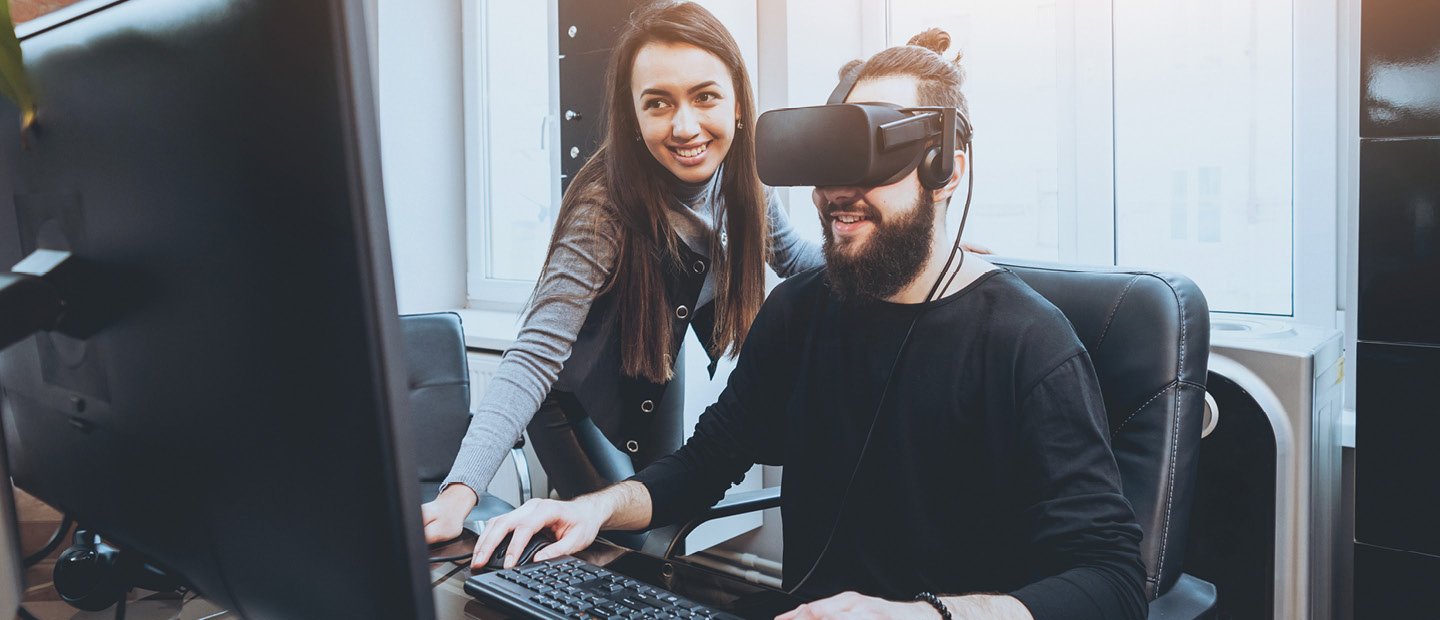man wearing a black virtual reality headset, seated at a computer with a woman standing next to him