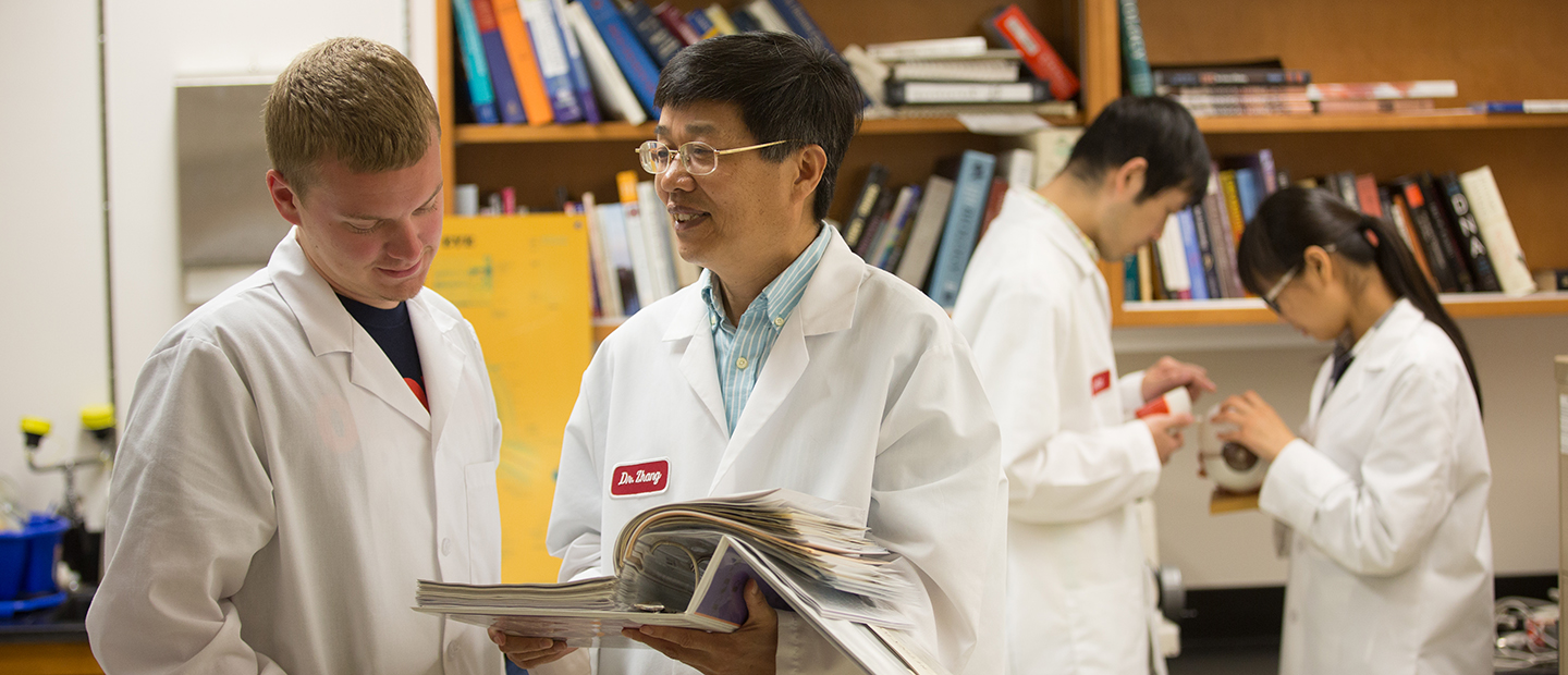 four people in white lab coats, standing in front of a book shelf, looking through a binder
