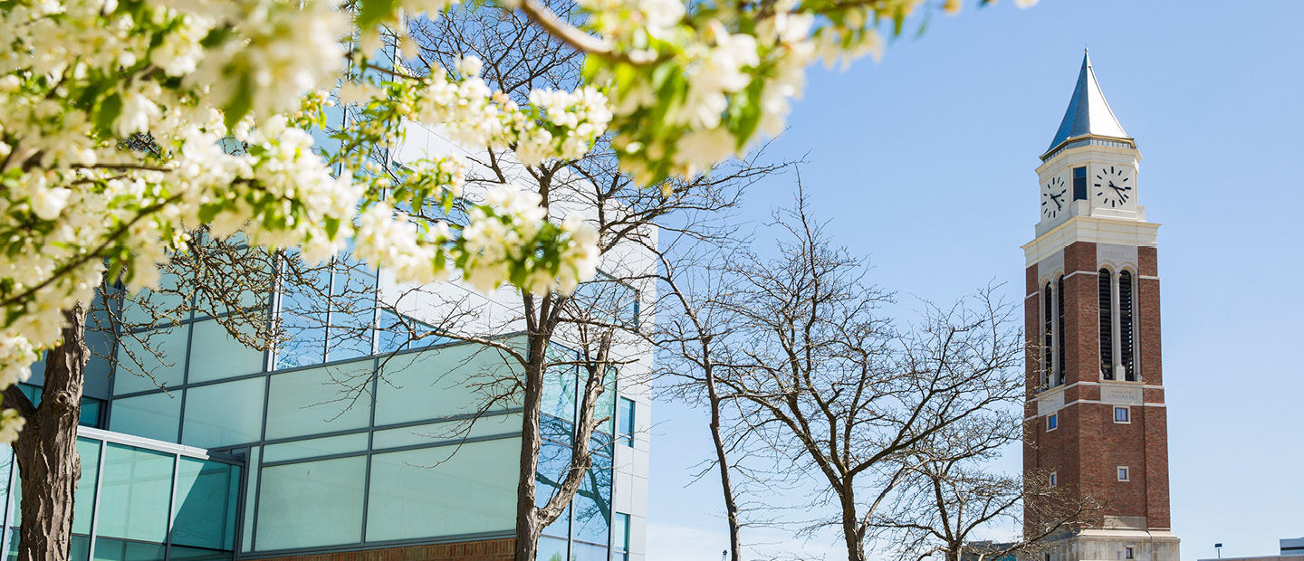 Elliott Tower and a blue glass building with a flowering tree in front on Oakland University's campus.