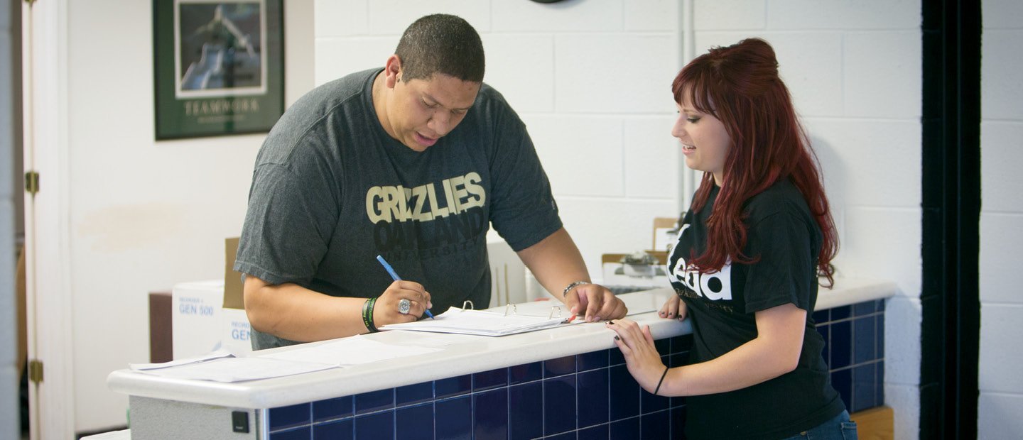 A man standing on one side of a partition, filling out paperwork, while a woman observes on the other side.