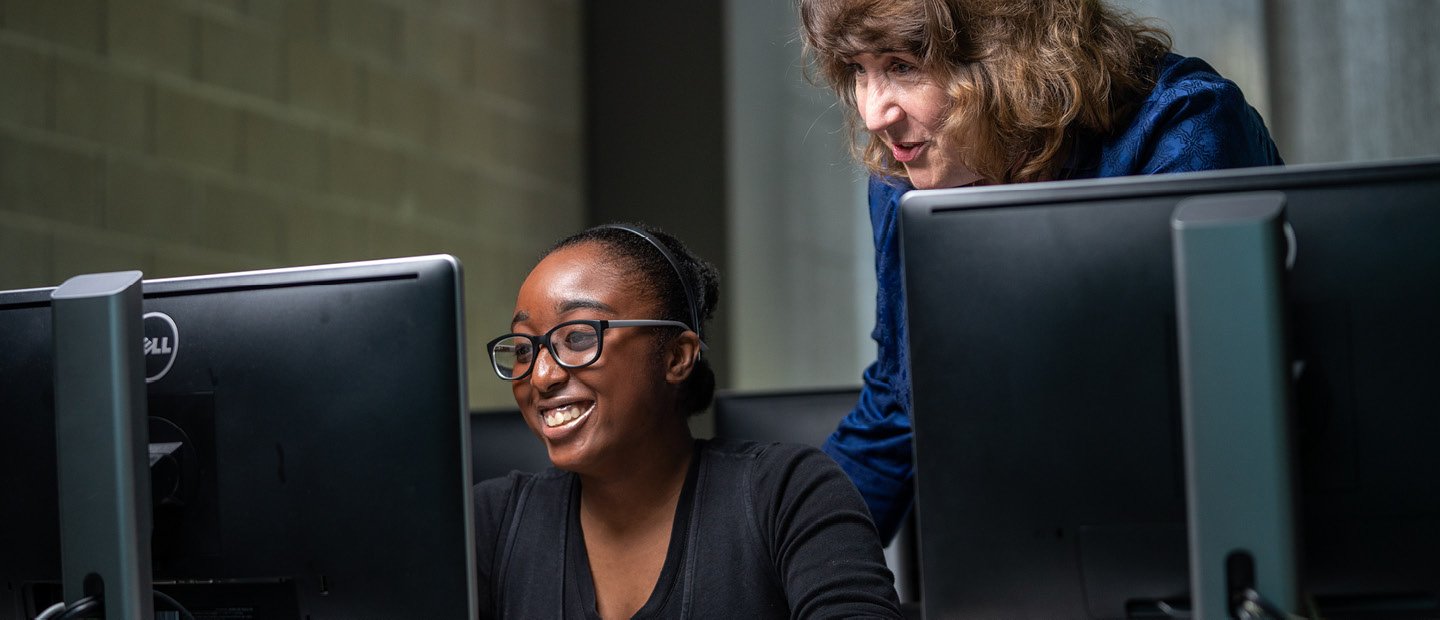 A young woman seated at a computer while a woman stands, looking over her shoulder.