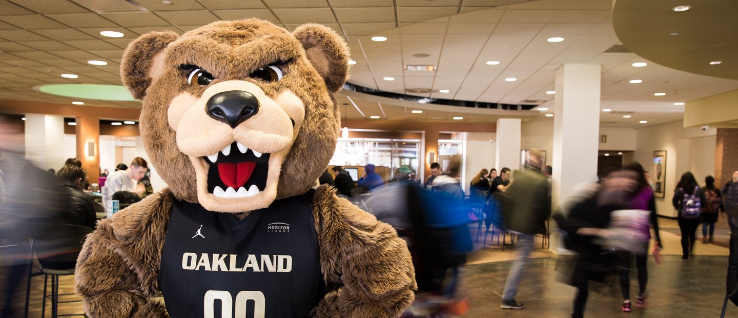 Apply_adm The Grizz bear wearing a jersey that says Oakland on it, with people walking through the room behind him