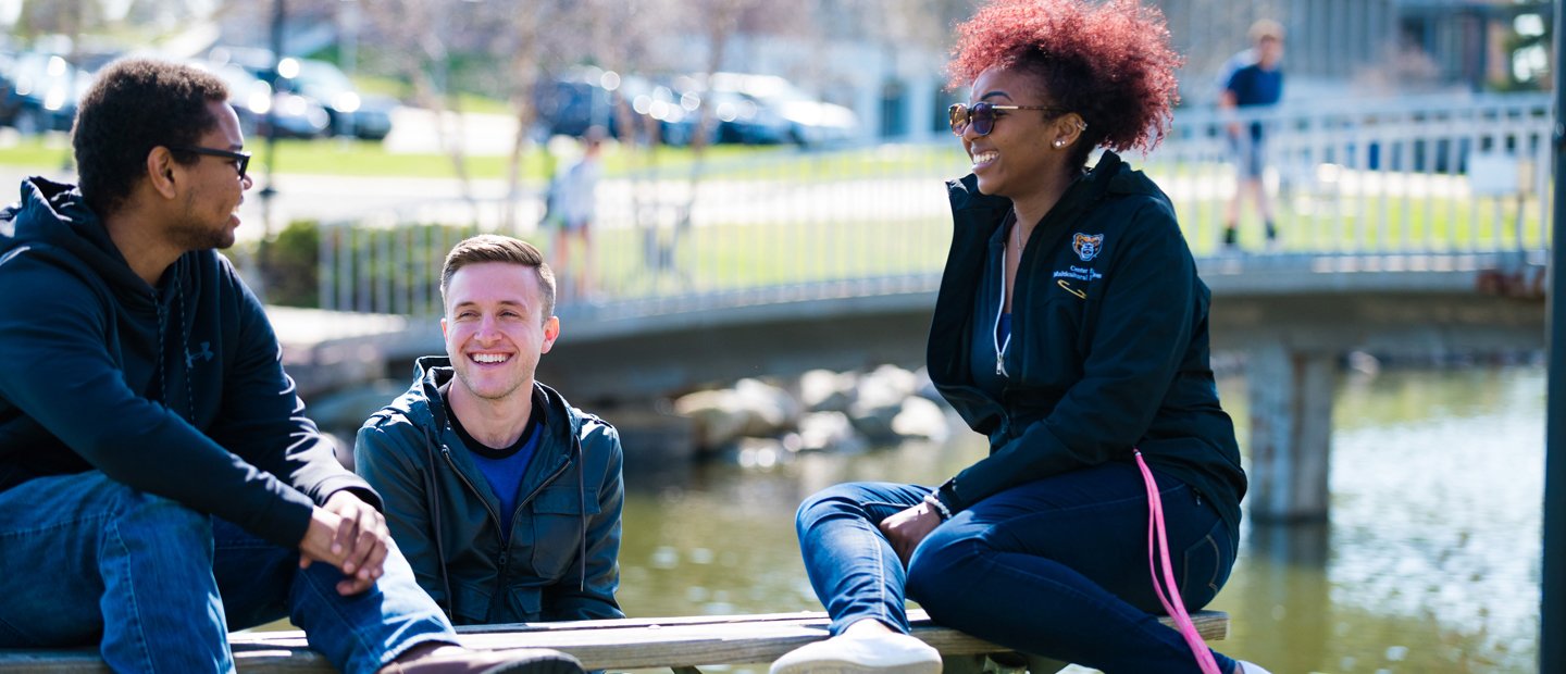 two male and one female O U student seated outside in front of a lake with a bridge in the background